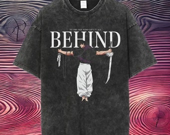 May include: Dark grey t-shirt featuring the word "BEHIND" and an illustration of a character holding a sword and a knife. The text "TO THE ONE WHO LEFT IT ALL" is above the word "BEHIND". The character is wearing a purple shirt and white trousers.