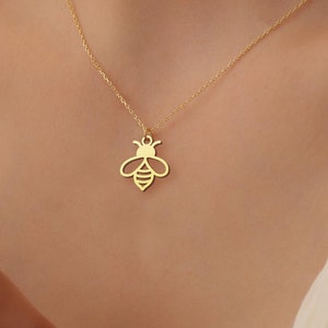 Puede incluir: Un collar dorado con un colgante de abeja. El colgante de abeja es un dise&ntilde;o recortado, que muestra el cuerpo y las alas del insecto. El collar se lleva alrededor del cuello, con la cadena visible contra la piel.