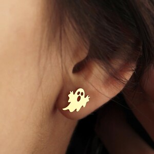 May include: Gold-colored ghost stud earrings. The earrings are small, with a simple ghost design. The ghost has a round body with a curved bottom and a small face with two eyes and a mouth. The earrings are worn on the ear.