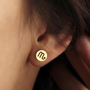 Peut inclure: Gros plan d'une boucle d'oreille dor&eacute;e avec un motif circulaire. La boucle d'oreille pr&eacute;sente un symbole stylis&eacute;, peut-&ecirc;tre un signe du zodiaque. La boucle d'oreille est sur l'oreille d'une personne, avec les cheveux et la main de la personne visibles en arri&egrave;re-plan.