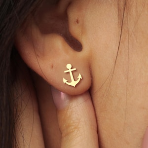 May include: A close-up of a gold-colored anchor stud earring. The earring is small and detailed, with a classic anchor design. The earring is worn on the ear. The background is blurred, focusing attention on the jewelry.