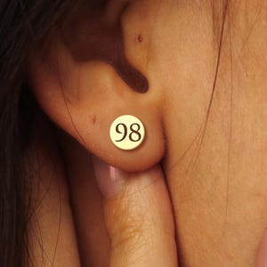 May include: A gold-coloured stud earring featuring the number "98" in a circular design. The earring is worn on an ear. The image shows a close-up of the ear and part of the face.