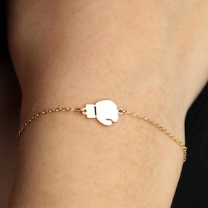 May include: A gold-coloured bracelet with a boxing glove charm. The charm is a solid, white silhouette of a boxing glove. The bracelet's chain is delicate and appears to be gold-plated. The bracelet is worn on a wrist.