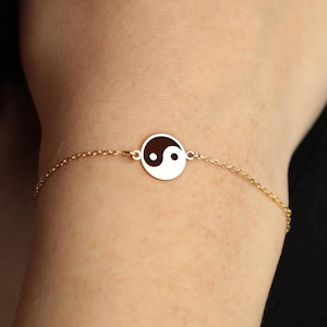 May include: Gold-colored bracelet featuring a circular pendant with a black and white yin yang symbol. The delicate chain is attached to the pendant, creating a minimalist and elegant design. The bracelet is shown on a person's wrist.