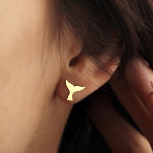 Puede incluir: Primer plano de una oreja con un pendiente de tuerca dorado con forma de cola de ballena. El pendiente es de dise&ntilde;o peque&ntilde;o y minimalista. La piel de la persona es clara. El fondo est&aacute; borroso.