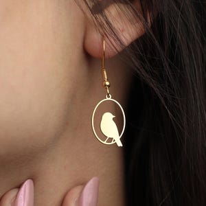 Puede incluir: Pendientes dorados con forma de p&aacute;jaro. Cada pendiente presenta una peque&ntilde;a silueta de p&aacute;jaro dentro de un marco circular, suspendida de un delicado gancho. Los pendientes se muestran en la oreja de una persona, destacando la elegante simplicidad del dise&ntilde;o y el tono c&aacute;lido del metal.