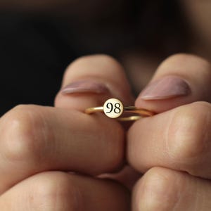 May include: A gold ring with a small, round, white face featuring the number 98 in black. The ring is held between two hands with manicured nails.