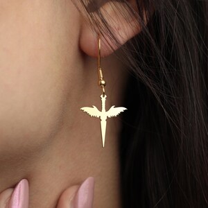 May include: A gold-coloured earring featuring a sword and wing design. The earring hangs from a delicate hook, with a central sword shape and wings on either side. The earring is worn on the ear, and the design is clearly visible.
