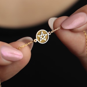 May include: A gold-colored bracelet featuring a circular pendant with a star design. The bracelet is held against a dark background, showcasing the delicate chain and clasp. The pendant has a light brown center.