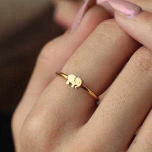 May include: A gold-colored ring featuring a small elephant design. The ring is thin and delicate, with the elephant charm centered on the band. The ring is worn on a finger, showcasing its simple elegance.