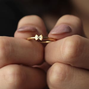 May include: A delicate gold ring featuring a small bow design. The ring is held between two hands, showcasing its intricate details. The bow is centered on the ring, adding a touch of elegance. The ring is a simple yet stylish piece of jewelry.