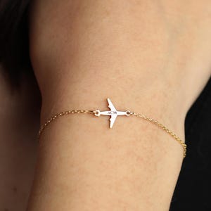 Könnte beinhalten: Ein zartes goldfarbenes Armband mit einem kleinen Flugzeug-Anhänger. Der Anhänger hat ein kreuzartiges Design mit einer kleinen Inschrift. Das Armband wird am Handgelenk getragen, eine passende goldene Halskette ist sichtbar.