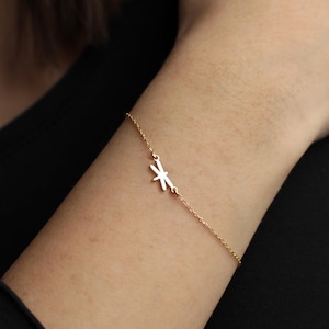 Könnte beinhalten: Ein filigranes goldenes Armband mit einem stilisierten Blumen- oder Sternmuster. Das Armband wird am Handgelenk getragen, mit einem schwarzen Oberteil im Hintergrund. Die Kette ist dünn und der Anhänger klein.