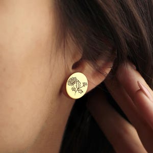 May include: Gold-colored oval stud earrings. Each earring features a detailed floral design. The earrings are shown on an ear, with a hand touching the ear. The earrings are a simple, elegant accessory.