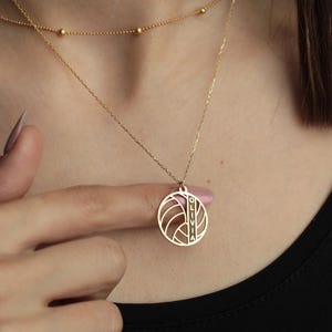 K&ouml;nnte beinhalten: Eine zarte goldene Halskette mit einem runden Anh&auml;nger mit geometrischem Design und dem eingravierten Namen "OLIVIA". Die Halskette ist mit einer zweiten, k&uuml;rzeren Kette mit kleinen goldenen Perlen versehen. Der Schmuck wird vor schwarzem Hintergrund pr&auml;sentiert.