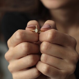 Pode incluir: Um anel dourado com um design de raquete de t&ecirc;nis. O anel &eacute; segurado entre duas m&atilde;os, mostrando a cabe&ccedil;a da raquete com um padr&atilde;o de grade. A banda do anel &eacute; fina e simples, complementando o design delicado da raquete. &Eacute; uma joia.