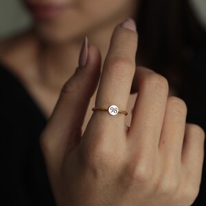 May include: A gold-coloured ring with a small, round, white charm. The charm features the number "98" in a simple, black font. The ring is worn on a finger, with the focus on the ring's design and details.