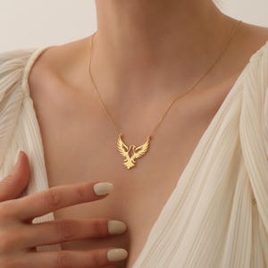 May include: A gold necklace with a delicate, openwork pendant shaped like a soaring bird. The pendant is a simple, elegant design with a minimalist aesthetic.