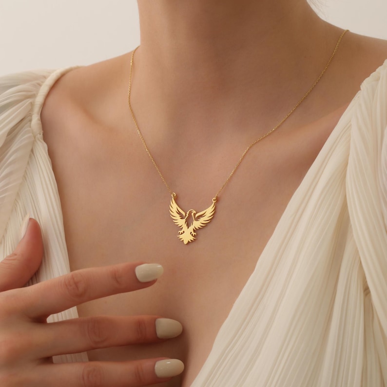 May include: A gold necklace with a delicate, openwork pendant shaped like a soaring bird. The pendant is a simple, elegant design with a minimalist aesthetic.
