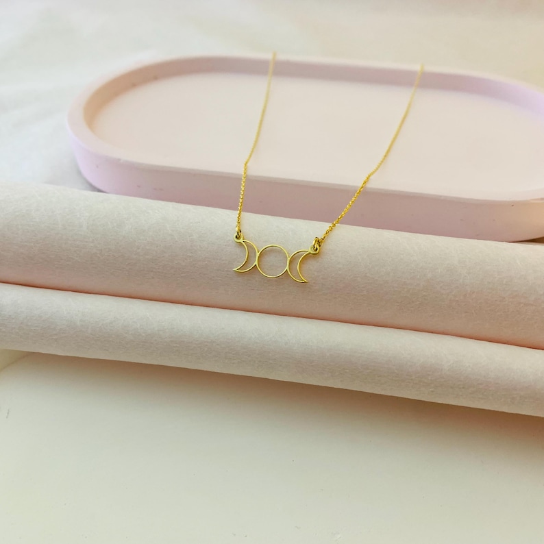 May include: A gold necklace with a triple moon pendant. The pendant is a simple design with three crescent moons.