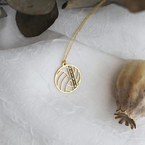 K&ouml;nnte beinhalten: Eine goldfarbene Halskette mit einem Volleyball-Anh&auml;nger. Der Anh&auml;nger hat ein ausgeschnittenes Design mit dem vertikal eingravierten Namen "OLIVIA". Die Halskette wird auf einer wei&szlig;en, strukturierten Oberfl&auml;che pr&auml;sentiert.