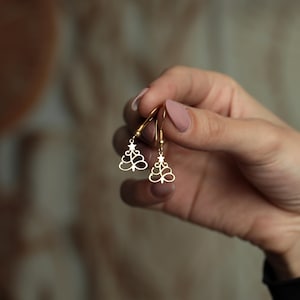 May include: A pair of gold-colored Christmas tree earrings. Each earring features a stylized tree design with a star at the top. The earrings are held up by a person's hand, showcasing the delicate details and festive theme.