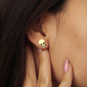 Puede incluir: Primer plano de un pendiente de bot&oacute;n de calavera dorada en una oreja. El pendiente tiene forma de calavera peque&ntilde;a y detallada con cuencas de los ojos y mand&iacute;bula. La piel de la persona es clara y se ve su pelo oscuro.