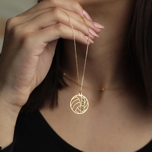 K&ouml;nnte beinhalten: Eine goldfarbene Halskette mit einem Anh&auml;nger mit einem Volleyball-Design. Der Anh&auml;nger tr&auml;gt den Namen "OLIVIA". Die Halskette hat eine zarte Kette.