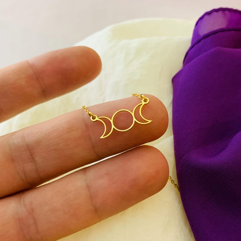 May include: A gold necklace featuring a triple moon pendant. The pendant is a simple design with three crescent moons and a circle in the centre.