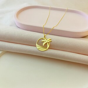 Peut inclure: Un collier dor&eacute; avec un pendentif repr&eacute;sentant un palmier et un oc&eacute;an dans un cercle. Le collier est pr&eacute;sent&eacute; sur un plateau rose et un tissu rose clair. La cha&icirc;ne est fine et le pendentif est d&eacute;taill&eacute;.