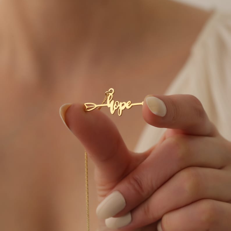 May include: A gold necklace with a delicate chain and a pendant featuring the word "hope" written in cursive script on an arrow.