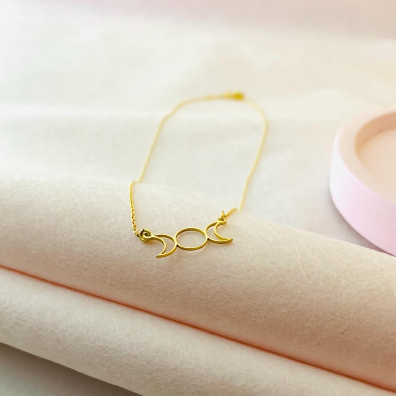 May include: A gold necklace featuring a triple moon pendant. The pendant is a simple design with three crescent moons.