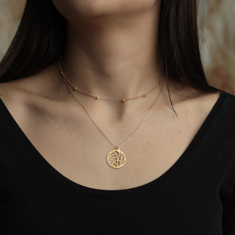 K&ouml;nnte beinhalten: Eine goldfarbene, mehrschichtige Halskette. Die obere Schicht ist eine zarte Kette mit kleinen, gleichm&auml;&szlig;ig verteilten Perlen. Die untere Schicht hat einen runden Anh&auml;nger mit geometrischem Design und einem vertikalen Streifen aus kleinen, dunklen Steinen. Die Halskette wird &uuml;ber einem schwarzen Oberteil getragen.