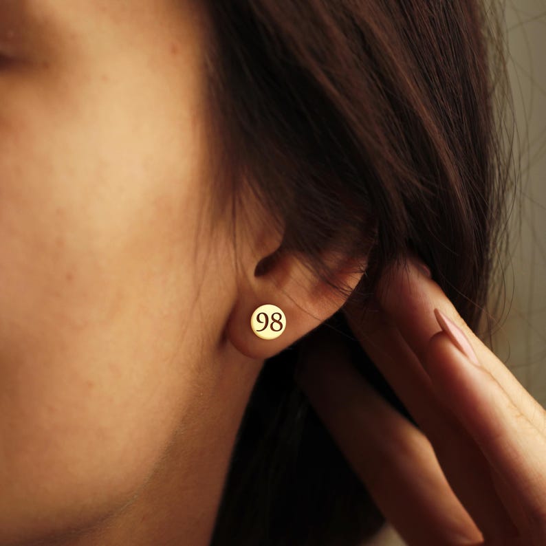 May include: A close-up shot of an ear with a gold stud earring. The earring is a small, circular disc with the number "98" printed on it. The skin is fair, and the hair is dark brown.