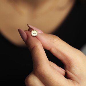 May include: A small, round gold-colored stud earring featuring a tennis ball design. The earring has a smooth, polished surface and a white tennis ball graphic. The earring is held between fingers against a dark background.