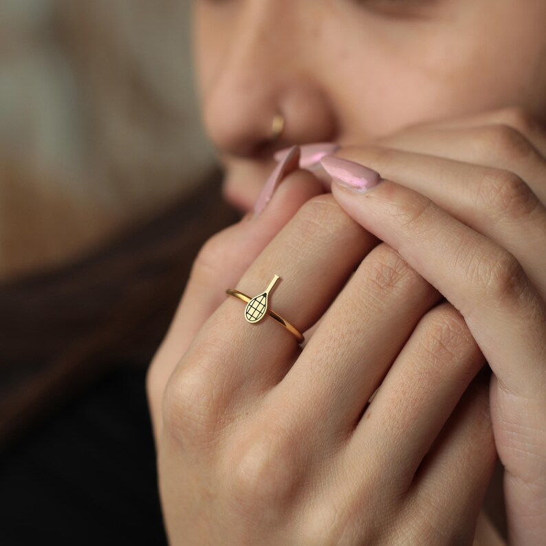 Pode incluir: Um anel dourado com um design de raquete de t&ecirc;nis. A cabe&ccedil;a da raquete tem um padr&atilde;o de grade. O anel &eacute; usado em um dedo. A banda &eacute; fina e simples. Uma joia.