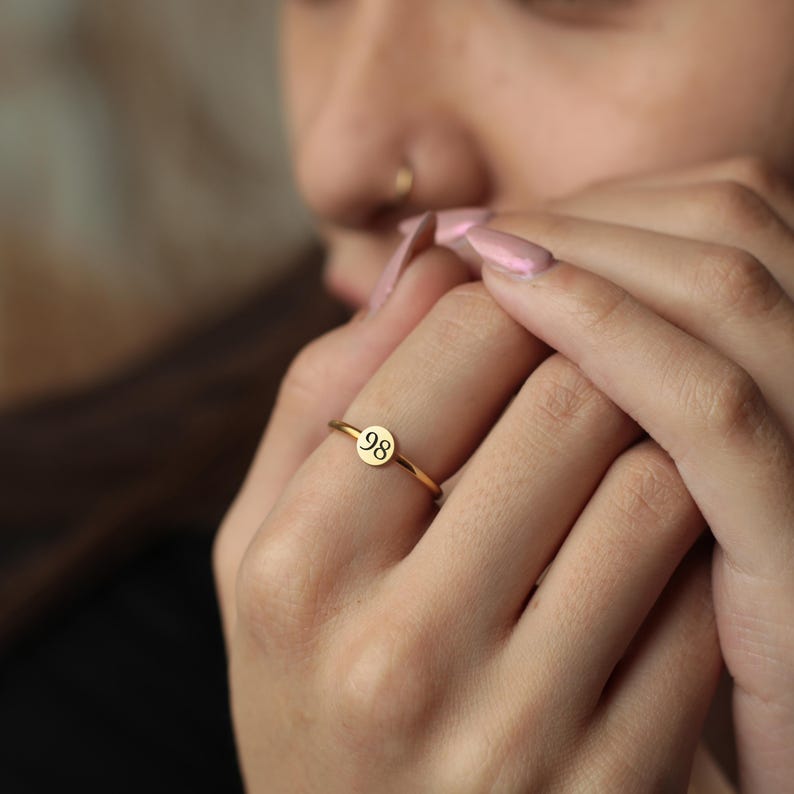 May include: A gold ring with a small, circular disc bearing the number "98". The ring has a slender band and is worn on a finger. The ring is a simple and elegant piece of jewellery.