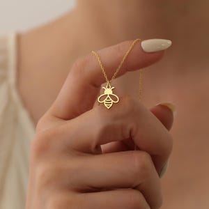 Puede incluir: Un collar dorado con un colgante de abeja. El colgante de abeja es una silueta estilizada con alas y segmentos corporales detallados. La cadena del collar es delicada y parece ser de oro. La joya se muestra sobre un fondo suave y neutro.