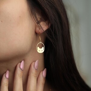 May include: Gold-colored cat-shaped dangle earrings. Each earring features a cat face silhouette within an oval frame, suspended from a simple hook. The earrings are shown on a person's ear, with a focus on the jewelry's design and color.