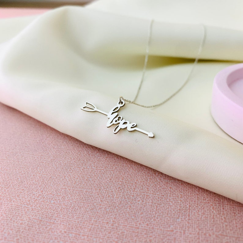 May include: Silver necklace with an arrow pendant featuring the word "hope" in cursive script.