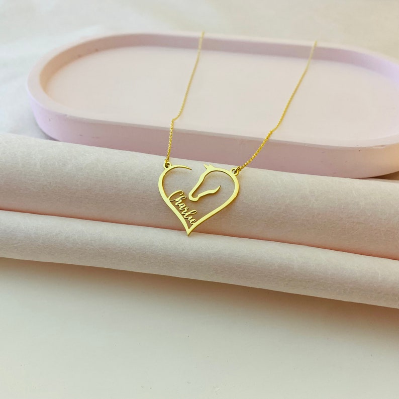 May include: Gold necklace with a horse head silhouette inside a heart shape. The name "Charlie" is inscribed on the pendant.