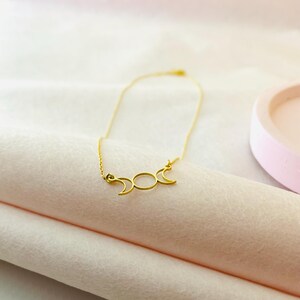 May include: A gold necklace featuring a triple moon pendant. The pendant is a delicate, open design with three crescent moons.
