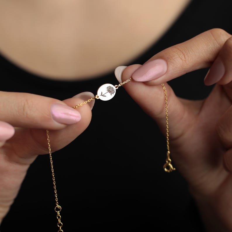 Op de afbeelding: Een delicate goudkleurige armband met een kleine, ronde hanger met een bloemmotief. De armband wordt tussen twee handen gehouden, waarbij de fijne ketting en sluiting te zien zijn. De hanger heeft een gedetailleerde gravure van een bloem.
