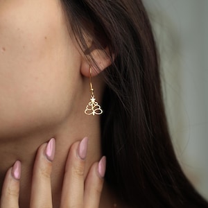 May include: Gold-colored Christmas tree earrings. The earrings feature a delicate tree design with a star on top, hanging from a simple hook. The earrings are shown on a person's ear, with their hand and hair in the frame.