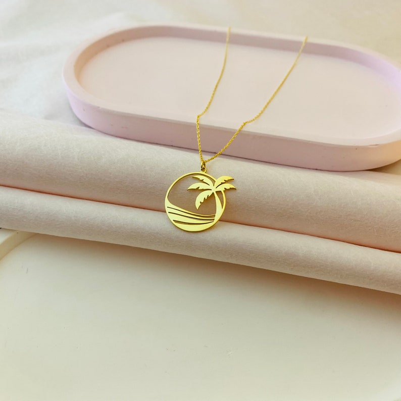 Puede incluir: Un collar de oro con un colgante de palmera y ola de mar. El colgante es un c&iacute;rculo con un dise&ntilde;o recortado de una palmera y olas del mar.