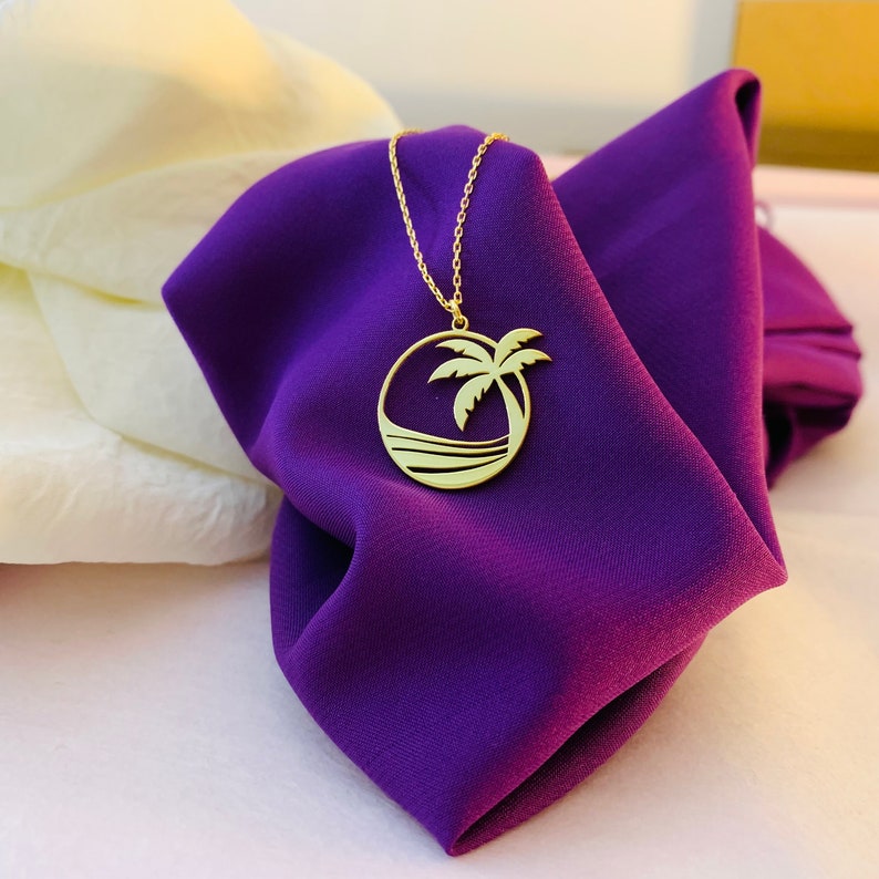 Peut inclure: Un collier dor&eacute; avec un pendentif repr&eacute;sentant un palmier dans un cercle. Le pendentif est suspendu &agrave; une fine cha&icirc;ne, pr&eacute;sent&eacute; sur un tissu violet fonc&eacute;. Le design comprend une sc&egrave;ne oc&eacute;anique stylis&eacute;e, &eacute;voquant un th&egrave;me tropical.