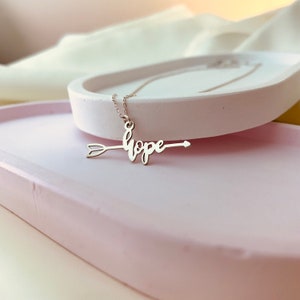 May include: Silver necklace with an arrow pendant featuring the word "hope" in cursive script.