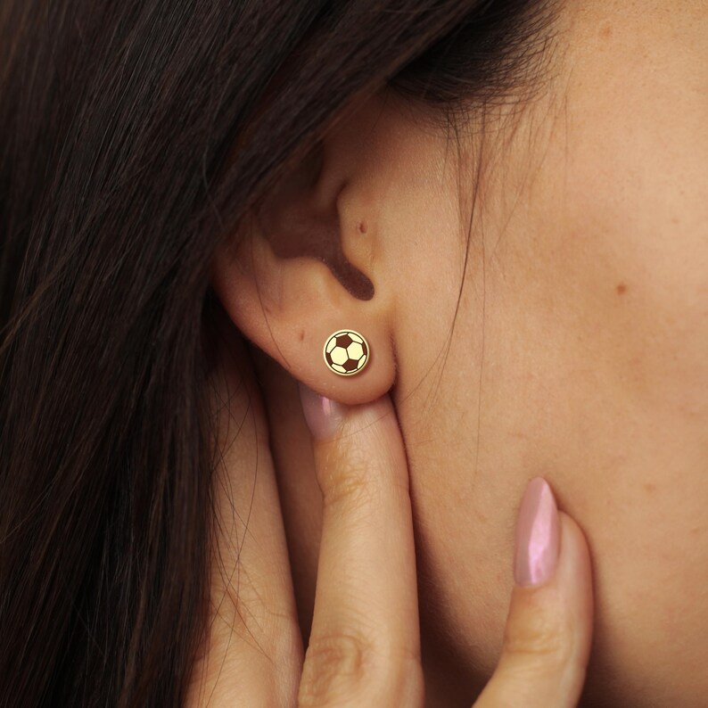 May include: A gold stud earring in the shape of a football, worn on a person's ear. The earring has black and white detailing. The person has fair skin and dark hair is visible in the background.