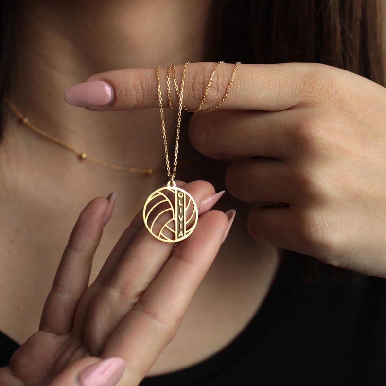 K&ouml;nnte beinhalten: Goldfarbene Halskette mit einem Anh&auml;nger in Form eines Volleyballs. Der Anh&auml;nger tr&auml;gt den Namen "OLIVIA" vertikal eingraviert. Die Halskette wird von den Fingern einer Person gehalten, wodurch die zarte Kette und die Anh&auml;ngerdetails hervorgehoben werden. Der Hintergrund ist unscharf.