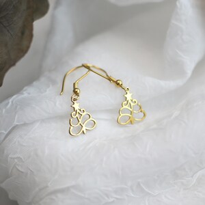 May include: A pair of gold-colored Christmas tree earrings. Each earring features a delicate, openwork design of a stylized tree with a star at the top. The earrings hang from simple, curved ear wires, set against a white, textured background.